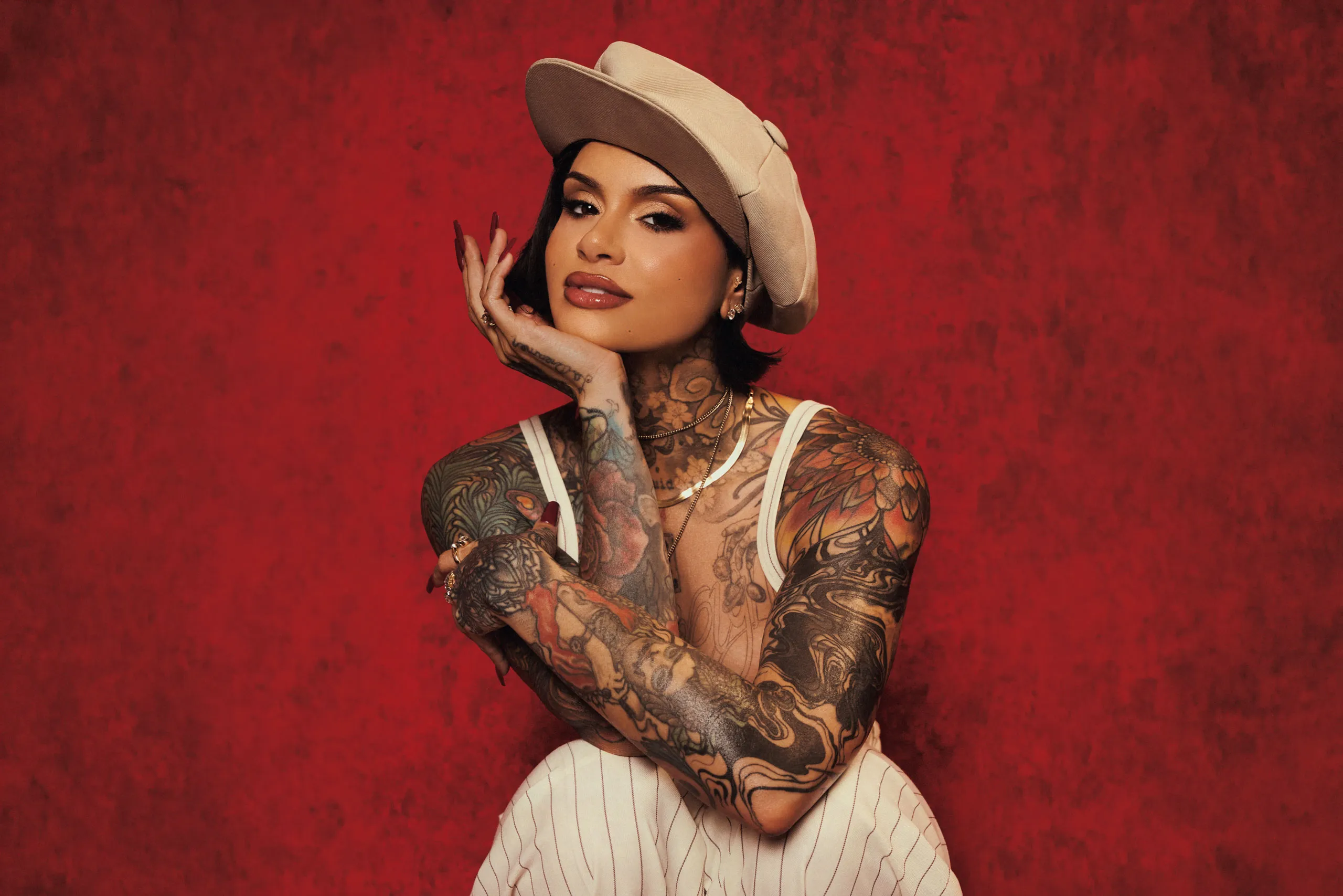 Fundo vermeho com a cantora Kehlani ao cento. A cantora é uma mulher negra com tatuagens cobrindo seu corpo até o pescoço. Ela esta usando uma roupa no tom de bege e listras verticais e uma boina de mesma cor.