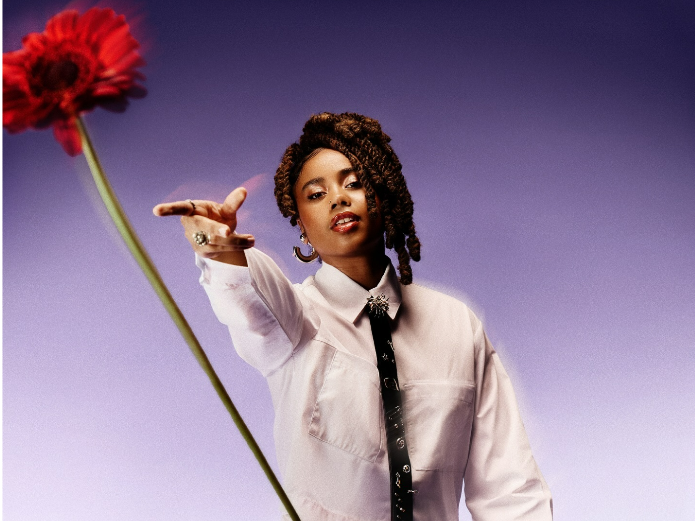 Cantora Melly, uma mulher negra com dreads castanho escuro com uma camiseta branca e gravta preta com alguns broches. A cantora esta com posição de estar jogando uma flor de cor vermelha e cabo verde