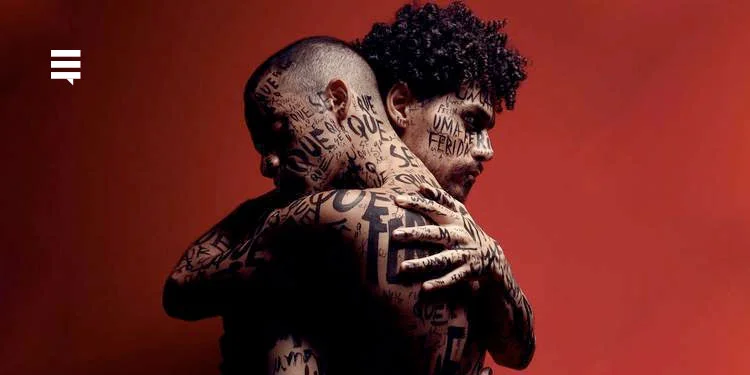 Fundo vermelho e dois homens se abraçando, ambos com varias palavras tatuadas espalhadas pelo corpo